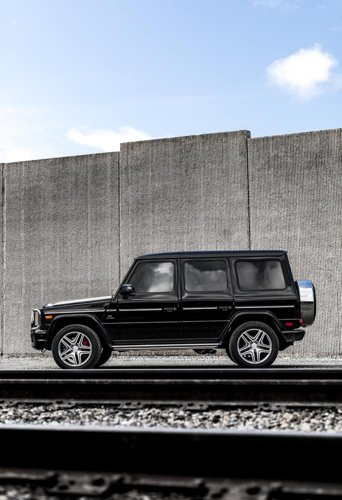 A black SUV with a ceramic coat and silver wheels is parked in front of a tall concrete wall. The sky is partly cloudy, providing a contrast to the vehicle's shiny exterior. Smooth asphalt and gravel are visible in the foreground.