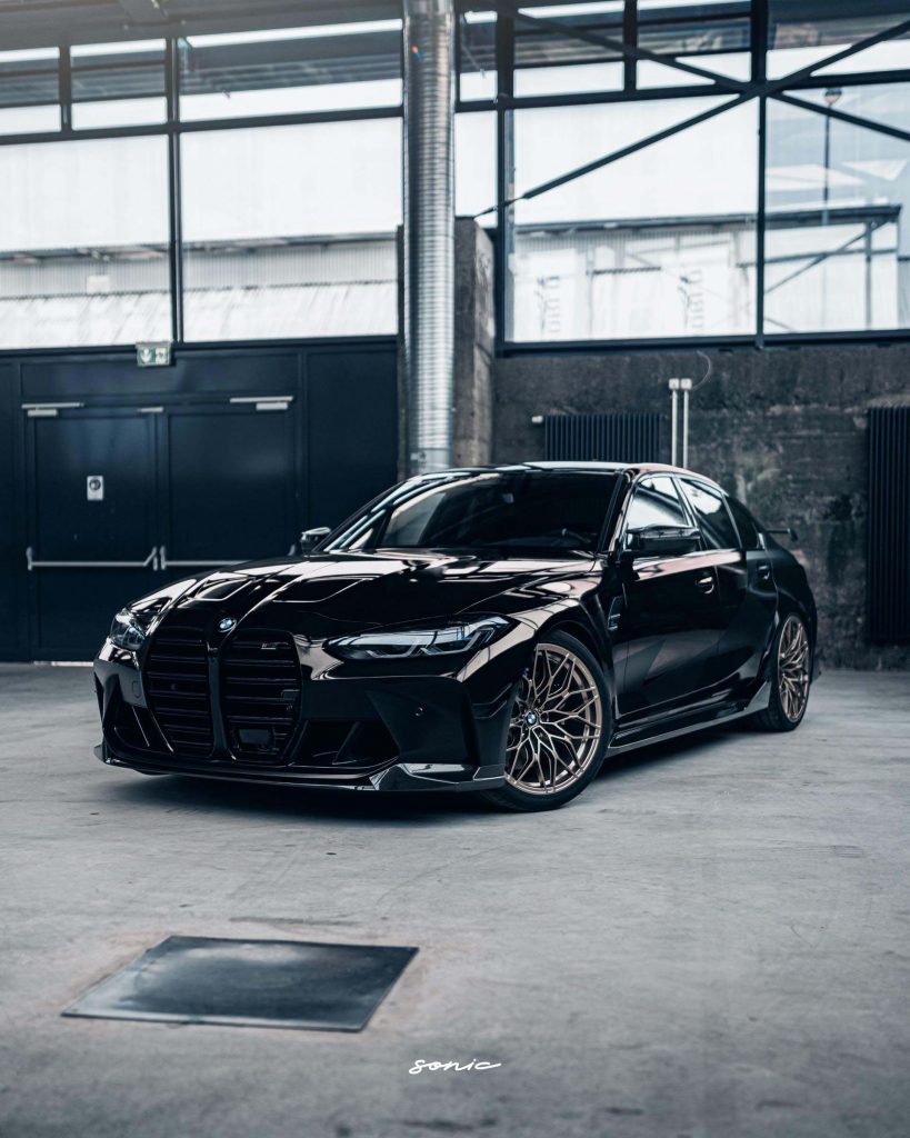 A sleek black luxury sports car is parked indoors with a modern, industrial backdrop. The car boasts a ceramic coat and stylish rims, highlighting its polished and high-performance design. Its interior is impeccably clean, reflecting meticulous auto detailing craftsmanship.