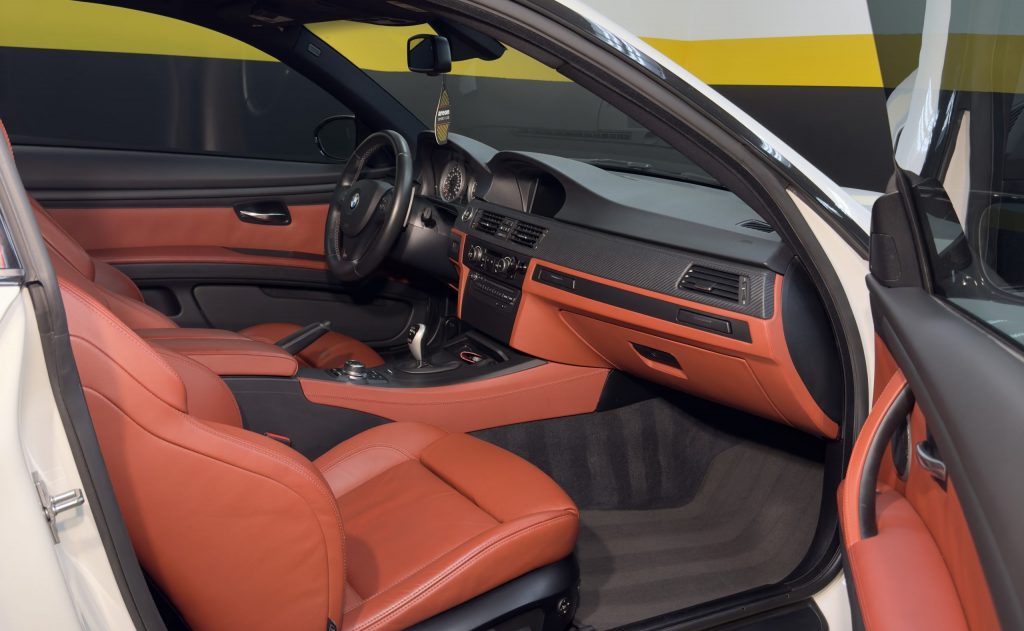 Interior of a modern car with orange leather seats and black accents, freshly enhanced by meticulous auto detailing. The dashboard and center console are sleek, featuring a digital display and various controls. The car is parked with the driver's door open, offering a pristine view inside.