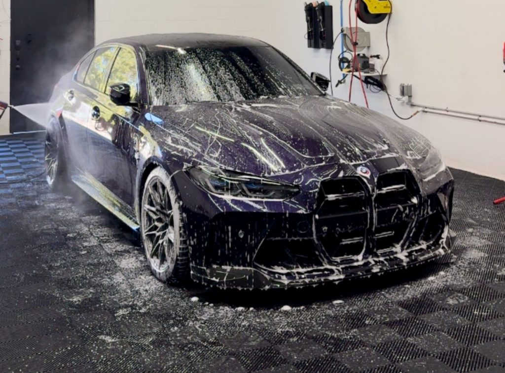 A dark-colored car undergoing auto detailing in a garage, covered with soap suds. It is parked on a black floor, with a hose and cleaning equipment visible on the wall in the background.