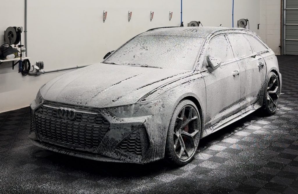 A silver Audi station wagon covered in white foam during a meticulous auto detailing session. It is parked indoors on a checkerboard-patterned floor, with wall-mounted hoses ready for precision work, enhancing its shine and protecting it with ceramic coat technology.